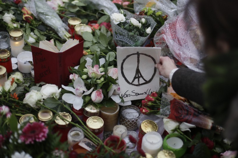 A woman places a paper with a peace sign combined with the Eiffel Tower and the words: ‘We are united’ between flowers and candles to remember the victims of Friday’s attacks in Paris, in front of the French Embassy in Berlin, Sunday, Nov. 15, 2015. Multiple attacks across Paris on Friday night have left scores dead and hundreds injured. CREDIT: AP PHOTO/MARKUS SCHREIBER