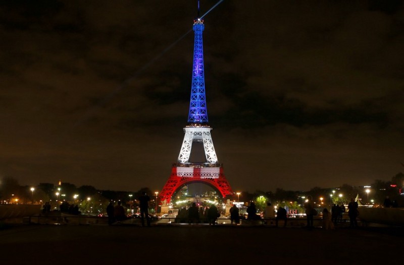 People look at the Eiffel Tower illuminated in the French colors in honor of the victims of the attacks on Friday in Paris, Monday, Nov. 16, 2015. CREDIT: AP Photo/Frank Augstein