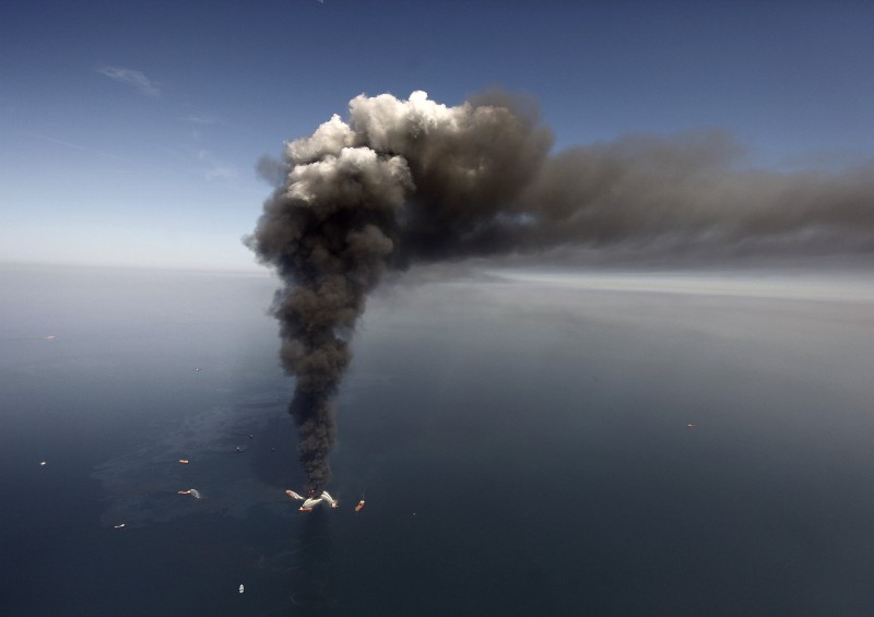 In this April 21, 2010 file photo, oil can be seen in the Gulf of Mexico, more than 50 miles southeast of Venice on Louisiana’s tip, as a large plume of smoke rises from fires on BP’s Deepwater Horizon offshore oil rig. (AP Photo/Gerald Herbert, File)