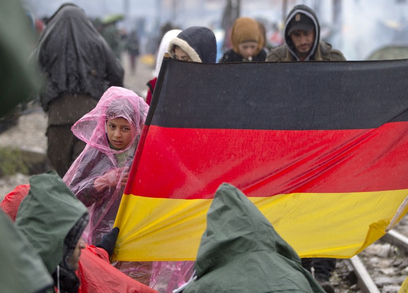 A child wearing a rain poncho stands next to a Germany flag as protesting migrants stage a sit in protest on the railway tracks at the northern Greek border station of Idomeni, Wednesday, March 9, 2016. Despair and confusion spread through the camp at the Greek-Macedonian border as thousands of stranded refugees were forced to acknowledge that the route through Europe that had carried their hopes and dreams was now shut. (AP Photo/Vadim Ghirda) CREDIT: AP PHOTO/VADIM GHIRDA