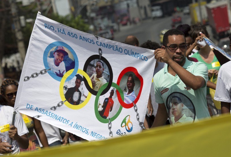 A man holds a banner designed with images of people killed during police operations against drug traffickers, framed in Olympic rings, with a message that reads in Portuguese; “”Rio, Olympic champion of the killing of Indians, and Black people for 450 years” during a protest at the Complexo de Alemao slum, in Rio de Janeiro, Brazil, Saturday, April 2, 2016. CREDIT: SILVIA IZQUIERDO, AP