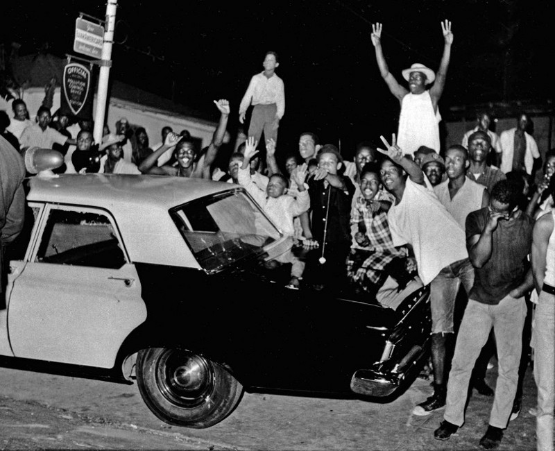 In this Aug. 12, 1965 file photo, demonstrators push against a police car after rioting erupted in the Watts district of Los Angeles. CREDIT: AP Photo