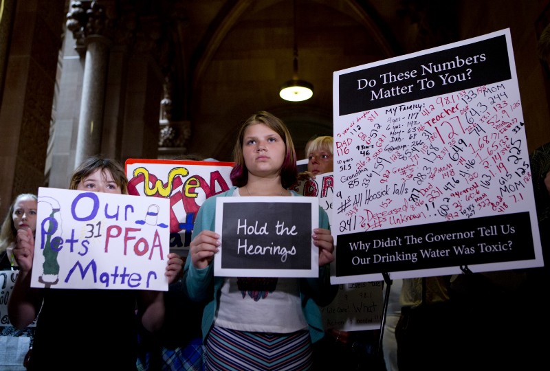 Hoosick Falls residents hold signs during a news conference at the state Capitol in Albany, N.Y., calling for hearings on the state’s handling of PFOA contamination in drinking water in their town. CREDIT: AP PHOTO/MIKE GROLL