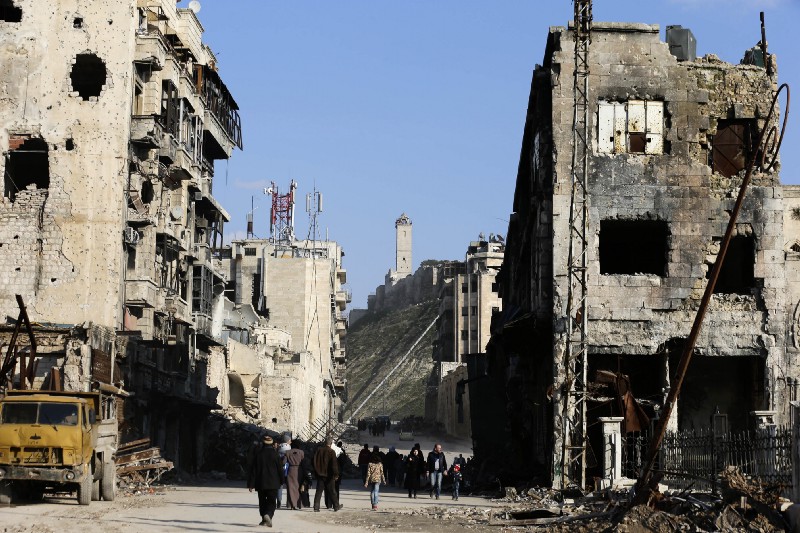Residents walk through damaged buildings near to the ancient Aleppo Citadel, background, that government troops used as a military base in the old city of Aleppo, Syria, Saturday, Jan. 21, 2017. CREDIT: AP Photo/Hassan Ammar