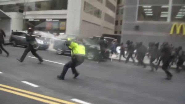 A masked, black-clad demonstrator jabs at an officer with a pole as the officer sprays him with a pepper cannon. CREDIT: YouTube/Screengrab