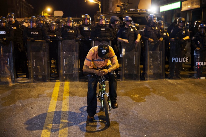 A man sits on a bicycle in front of a line of police officers in riot gear ahead of a 10p.m. curfew in the wake of riots following the funeral for Freddie Gray, April 2015, in Baltimore. CREDIT: AP Photo/David Goldman