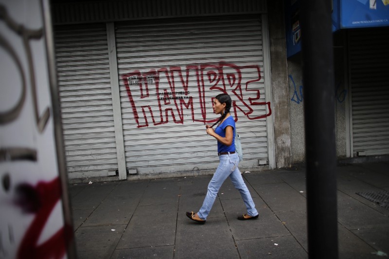 A woman walks past a shuttered shopfront with “hunger” painted on it, in Caracas, Venezuela. Large swaths of Venezuela’s capital were shuttered and silent Thursday as opponents of President Nicolas Maduro called a major national strike. CREDIT: AP Photo/Ariana Cubillos