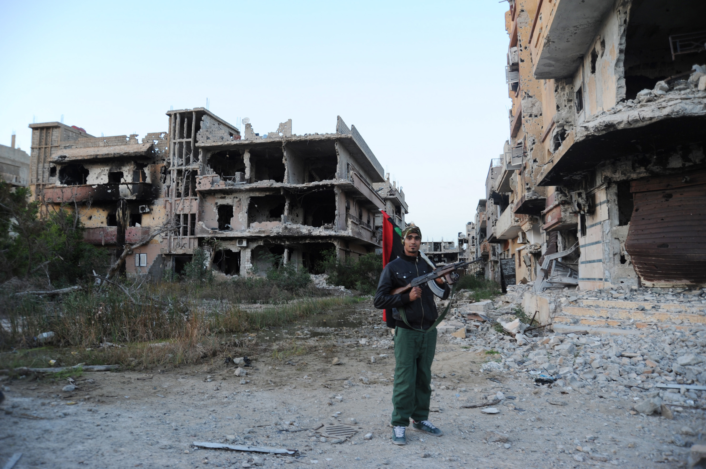 A civilian fighter holding the Libyan flag stands in front of damaged buildings in Benghazi, Libya. The U.S., Europe and U.N. have all pinned their hopes for resolving Libya's chaos and blocking the Islamic State group's growth there on a newly announced unity government. Headed by a little known Libyan technocrat, Fayez Serraj, it is supposed to replace the two rival administrations - one based in the capital Tripoli, the other based in the eastern city of Tobruk- that have been battling each other. CREDIT: Mohammed el-Shaiky/AP Photo.