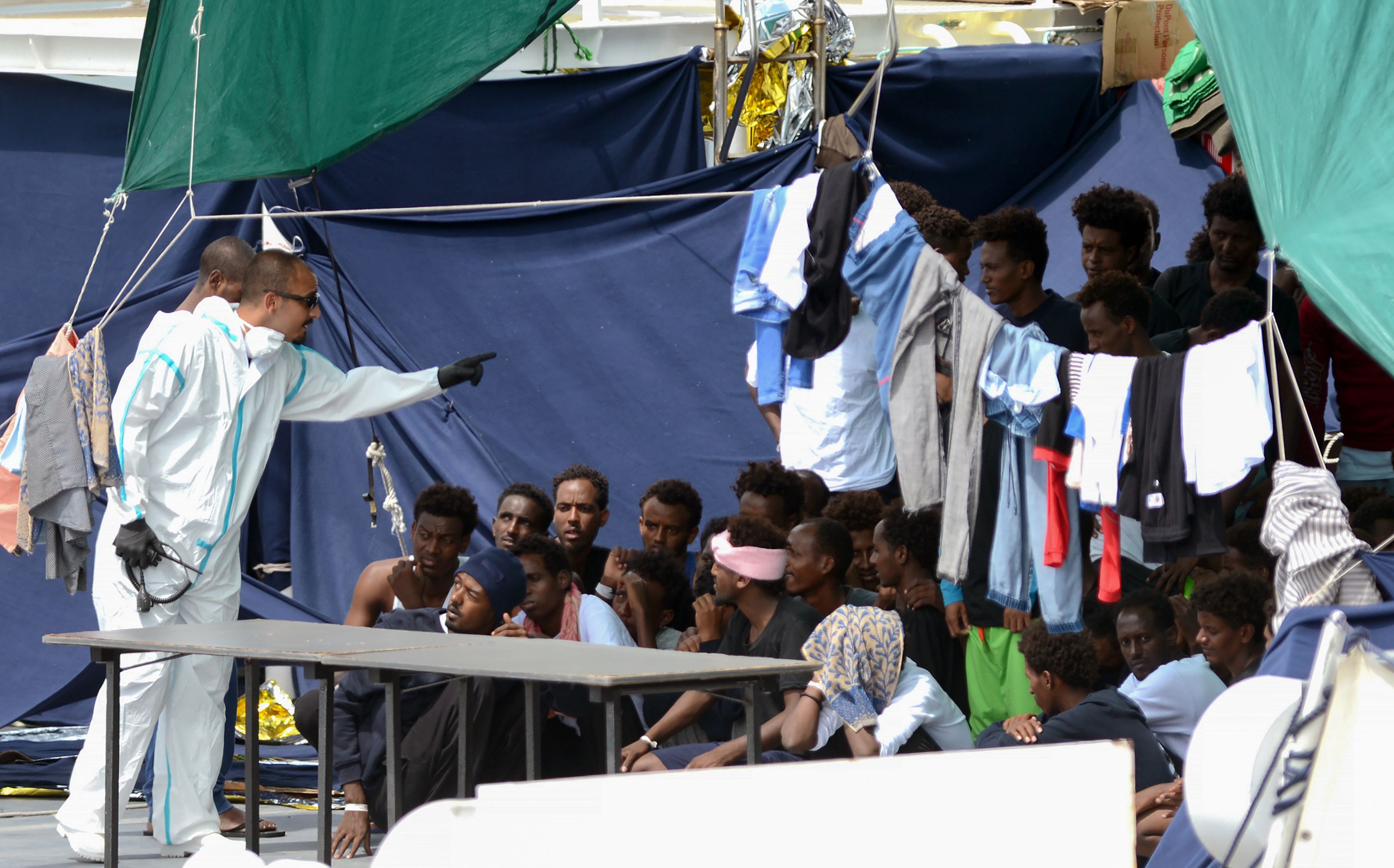 An official wearing a protective suit gestures towards migrants as they sit on the deck of the Italian Coast Guard vessel "Diciotti" in the Sicilian port of Catania, on August 23, 2018, as they wait to disembark following a rescue operation at sea. CREDIT: Giovanni Isolino/AFP/Getty Images.