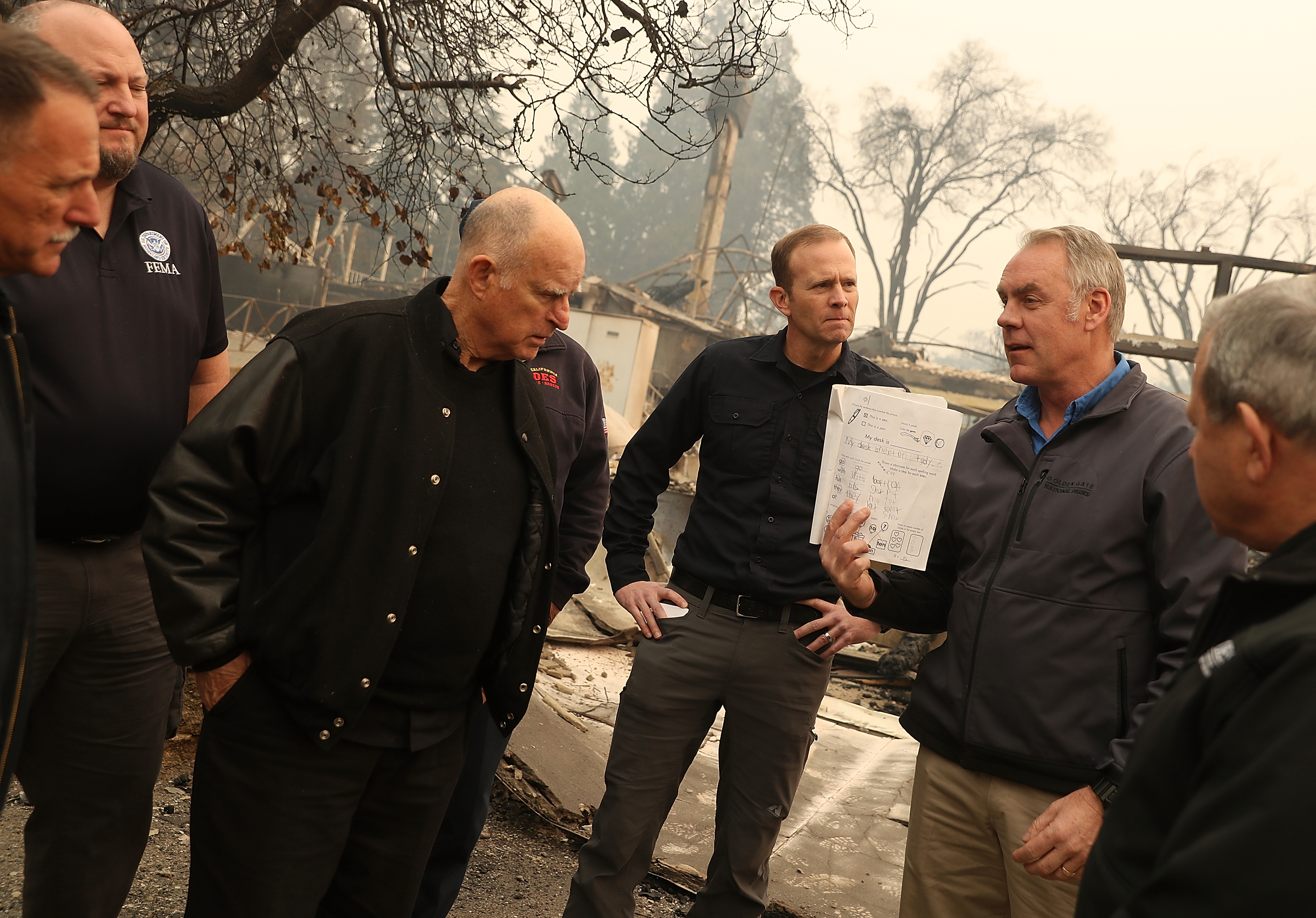 California Governor Jerry Brown and FEMA Adminstrator Brock Long look on as U.S. Secretary of the Interior Ryan Zinke holds up a child's school work as they tour a school burned by the Camp Fire on November 14, 2018 in Paradise, California. CREDIT: Justin Sullivan/Getty Images